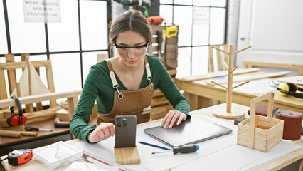 A woman uses a smartphone in a bright woodworking studio filled with tools and wooden items.