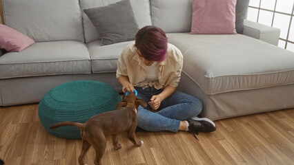 A young caucasian woman sits on the floor in her living room playing with her dog, creating a warm and cozy indoor scene.