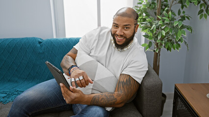A smiling bearded black man lounges comfortably with a tablet in a modern apartment living room.