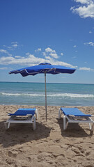 A serene beach scene with an open blue umbrella, white sunbeds, golden sand, clear sky, and a tranquil sea.