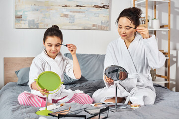 A brunette woman brushes her hair while her daughter sits on a bed, both dressed in white bath robes, applying makeup