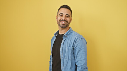 Handsome young hispanic man with a beard smiling against a yellow wall background.