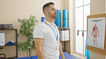 Young hispanic man with beard, standing thoughtfully in a rehab clinic indoors, exuding professionalism and care.