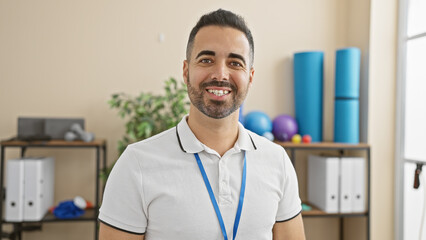 Handsome hispanic man with beard smiling in a clinic rehab indoor setting wearing a lanyard
