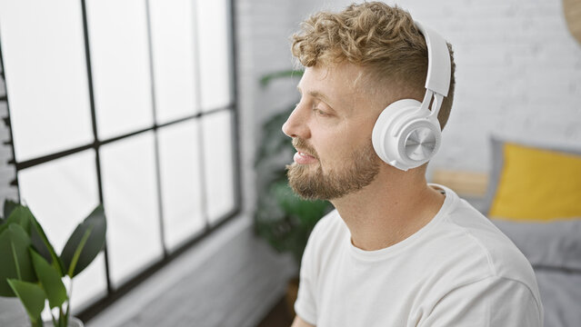 A young caucasian man with blue eyes and a beard enjoys music on headphones in a cozy bedroom setting