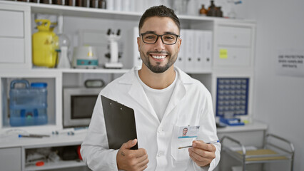 Smiling man with beard wearing glasses posing in a lab coat holding a clipboard and id card in a...
