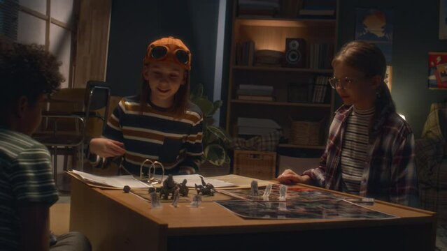 Handheld shot of Caucasian boy explaining rules of board game to diverse friends who looking at cards and maps at night