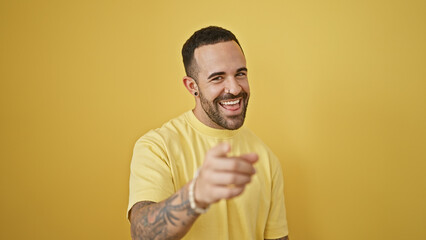 A cheerful young hispanic man with a beard, smiling and pointing, isolated against a yellow wall background.