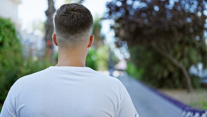 A young man in a white shirt walking outside on a city street surrounded by green foliage.