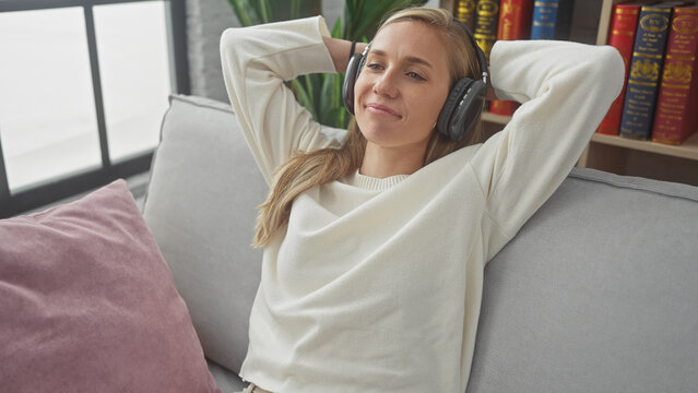 Relaxed young woman with blonde hair enjoying music on headphones while reclining on a sofa at home.