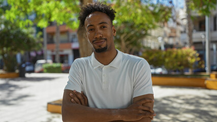 Fototapeta premium A handsome young african american man with a beard stands outdoors in an urban park setting on a sunny day, confidently crossing his arms in a city environment.
