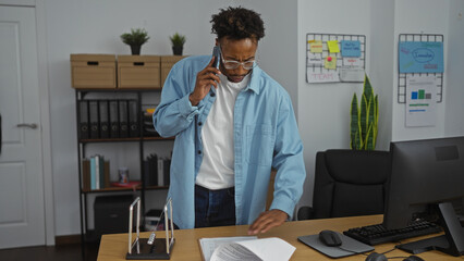 Young african american man with a beard talking on the phone while working at his desk in a modern office setting.
