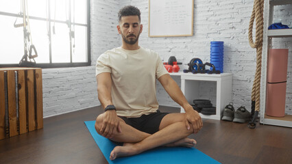 Young hispanic man meditating indoors in a gym, sitting cross-legged on a blue mat with gym...