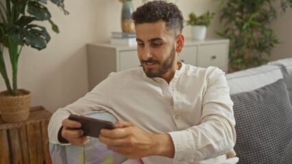 Handsome young hispanic man with a beard sitting on a couch in a cozy living room at home, looking at his phone.