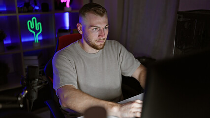 A focused young man with a beard works on a computer in a dark, neon-lit gaming room at night.