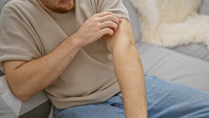 A bearded young caucasian man scratches his arm while sitting on a sofa indoors, evoking a casual, relaxed atmosphere.