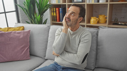 Obraz premium A thoughtful mature man with a beard sits contemplatively on a sofa in a cozy living room setting.