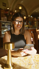 Smiling young woman with glasses using smartphone at a brightly lit cafe table