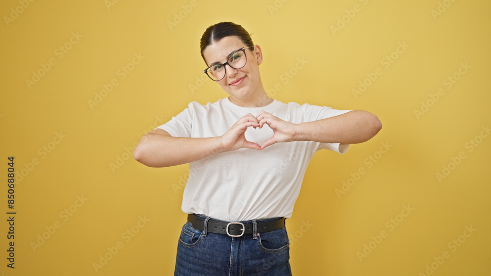 Sticker Young beautiful hispanic woman smiling confident doing heart gesture with hands over isolated yellow background