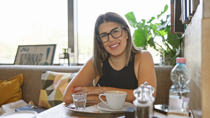 A cheerful young hispanic woman wearing glasses sitting in a bright indoor cafe with a coffee cup on the table.