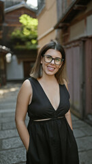 Confident hispanic woman, beautiful and joyful, cheerfully posing with glasses on, standing and smiling on kyoto's traditional streets, radiating natural and carefree latin expression.
