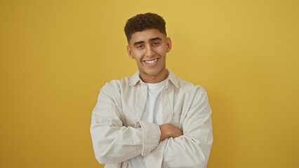 A smiling young adult man with crossed arms wearing a white shirt against a yellow background.