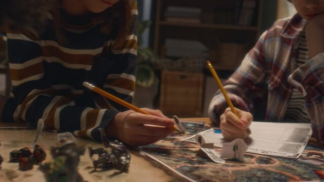 Midsection panning shot of unrecognizable diverse kids writing info into character sheets using cards and action figures before playing table game at home in evening