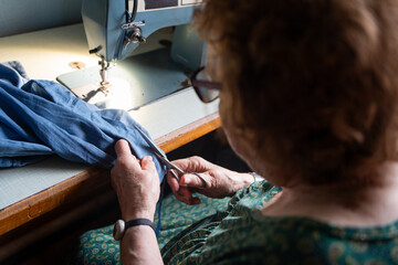 chubby eighty-year-old woman in green summer dressing gown mending and sewing the bottom of a pair...