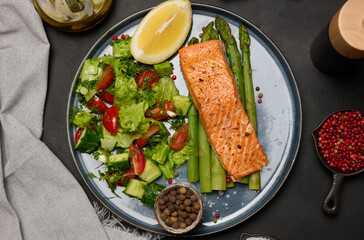 Healthy lunch with grilled salmon with asparagus and fresh tomato and cucumber salad, a row of lemon slice.