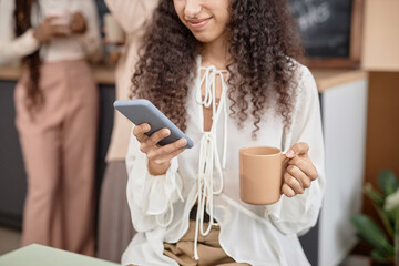 Cropped close up of Middle Eastern woman holding smartphone in blue case and beige cup and smiling in office or classroom, copy space