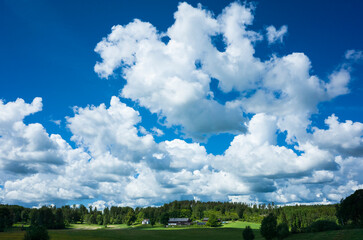 Cloudscape with white fluffy clouds on blue sky over Scandinavian rural landscape, Nature of Sweden in summer, Farmhouses by forest and green fields