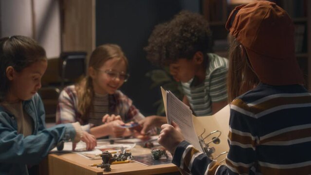 Back shot of boy writing down info about process of playing board game while his diverse friends discussing their character cards at home in evening