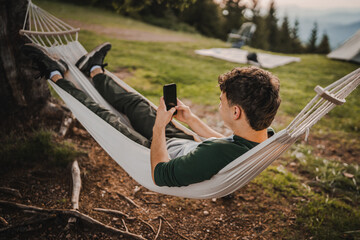 Young boy lying down in hammock and use mobile phone on the mountain