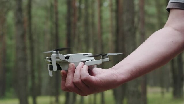 A quadcopter operator with a remote control holds the drone in front of him in focus outdoors. The drone takes off.