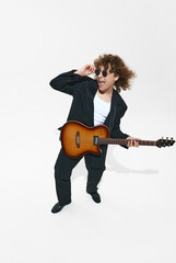 Musician in sunglasses and a suit, young man playing guitar, making creative performance isolated on white background. Concept of music, festival, concert, hobby, talent, performance