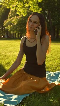 Front view of a female sitting towards camera talking during a phone call outdoors in the park. Slow motion shot. Vertical shot.