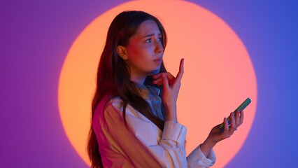 Stylish young woman in casual clothing holds smartphone, hesitates sending text message, confused...