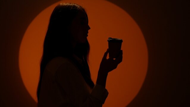 Stylish young woman silhouette in casual clothing drinking coffee from paper cup . Isolated on dark studio background in spotlight.