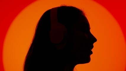 Stylish young woman black silhouette listening music in headphones, face close up. Isolated on red studio background spotlight.