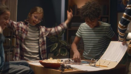 Side shot of Caucasian boy bringing potato chips and popcorn to group of diverse friends during snack time in board game at home in evening