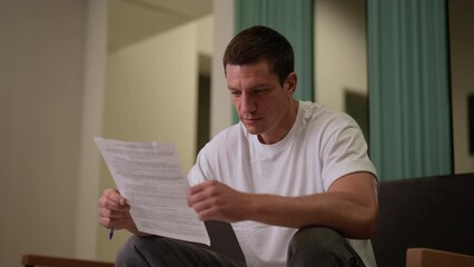 Focused male patient in casual clothes reading paper medical contract before signing and having medical procedure performed at hospital, while sitting on sofa in waiting room. Shooting in slow motion.
