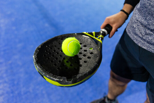 Man a holding a padel racket and and a yellow padel ball in a blue court 
