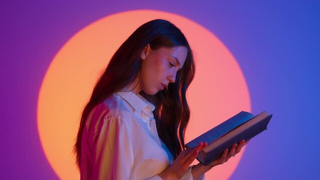Stylish young woman student in casual clothing holding and reading a book. Isolated on colorful studio background spotlight.