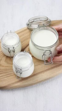 Homemade natural yoghurt, fermented in jars. Female hands twist an open jar of yogurt on a wooden board on light gray