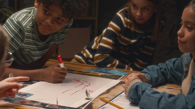 Side shot of group of multiethnic kids creating location map together for board game at home in evening, boy drawing with red marker pen