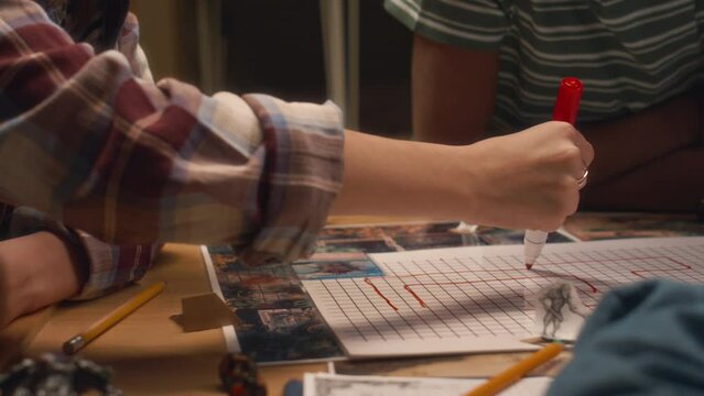 Side tilt shot of girl in glasses drawing lines in location map using red marker pen before playing tabletop game with friends at home in evening