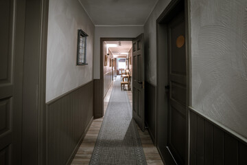 A cozy hotel corridor with open doors, a patterned rug, and elegant wooden furniture at the end. Soft lighting and warm tones create a welcoming atmosphere.
