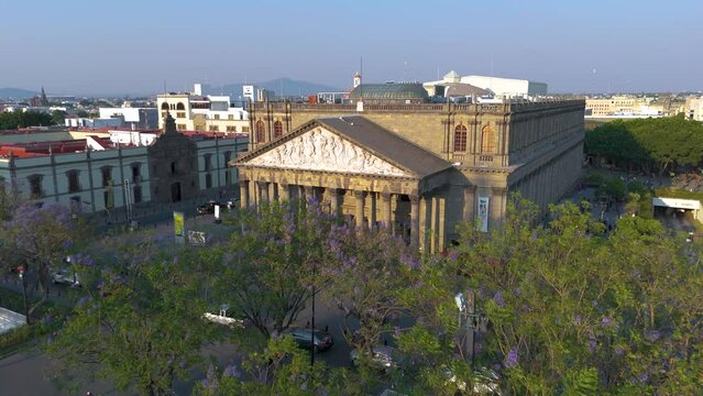 Teatro Degollado shows its facade warmly lit by the afternoon sun. Locals joyfully stroll around the theater, enjoying the plaza adorned with blooming purple Jacaranda trees.