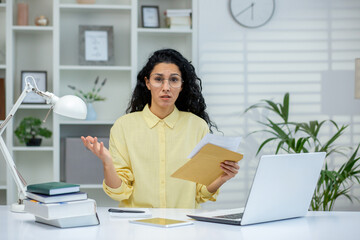 Portrait of young worried hispanic woman sitting at desk in modern bright office, received letter...