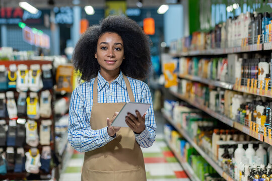 Portrait of a smiling African-American female consultant and assistant wearing a scarf standing in a supermarket among shelves with household chemicals, holding a tablet and looking at the camera.